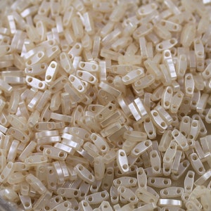 Può includere: Un grande mucchio di piccole perline ovali di colore bianco sporco con due fori in ogni perlina. Le perline sono in plastica e hanno una finitura leggermente iridescente.