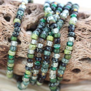 May include: A close-up of a strand of green, silver, and black seed beads. The beads are arranged in a random pattern and are strung on a clear thread.