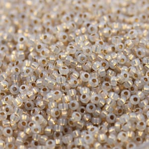 May include: A close-up of a pile of small, white, iridescent glass beads with gold-colored rims. The beads are uniform in size and shape, and they are arranged in a random pattern.
