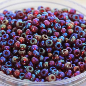 May include: A close-up of a bowl filled with small, iridescent glass beads. The beads are a deep red color with a blue and green iridescent sheen.