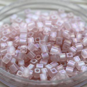 Könnte beinhalten: Eine Ansammlung kleiner, blassrosa würfelförmiger Perlen mit irisierendem Finish. Jede Perle hat ein mittiges Loch und sie befinden sich in einer runden, durchsichtigen Kunststoffschale, was ein einheitliches, reflektierendes Bild ergibt.