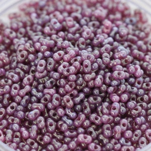Puede incluir: Primer plano de una gran cantidad de pequeñas cuentas de vidrio púrpura. Las cuentas tienen forma de ocho y un acabado brillante e iridiscente.