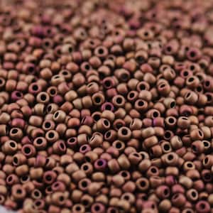 May include: Close-up of a pile of small, round beads in shades of brown, bronze, and hints of purple. The beads have a matte finish and are densely packed together, creating a textured surface. Ideal for jewelry making and crafts.