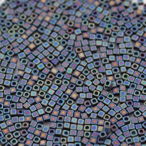 May include: A close-up view of a large quantity of iridescent, square-shaped beads. The beads are a dark blue color with hints of purple and gold.