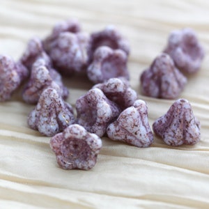 May include: A collection of small, white glass beads with a purple and gold speckled pattern. The beads are shaped like a flower with five petals.