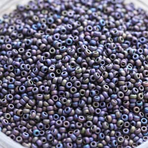 May include: A close-up shot of a pile of small, iridescent purple seed beads. The beads have a matte finish and a hole running through the center. The color shifts between purple, blue, and green, creating a shimmering effect. These beads are ideal for jewelry making and crafts.