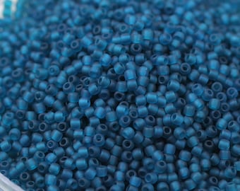 May include: A close-up view of a large quantity of small, opaque, teal blue glass beads. The beads are round and have a slightly frosted finish.