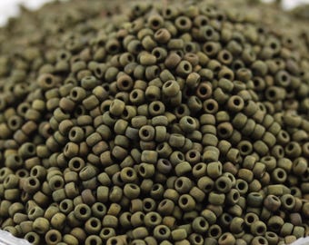 May include: A close-up shot of a pile of small, olive-green seed beads. The beads are cylindrical with a hole running through the center, perfect for stringing. The beads have a matte finish and a slightly irregular shape, suggesting a handmade quality.