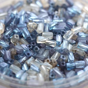 May include: A close-up of a variety of small, rectangular glass beads in shades of blue, clear, and white. The beads are scattered in a pile, creating a shimmering effect.