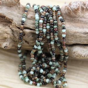 May include: A strand of seed beads in shades of brown, black, and teal green. The beads are arranged in a loose pile on a piece of driftwood.