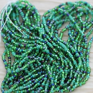 May include: A collection of green beaded strands. The beads vary in shade, from light to dark green, with hints of iridescent blue and brown. The beads are strung together, creating a textured and layered effect. White string is visible at the top.