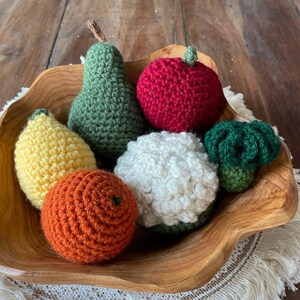 May include: A wooden bowl filled with crocheted fruit and vegetables, including a green pear, a yellow lemon, an orange orange, a red apple, a white cauliflower, and a green broccoli.