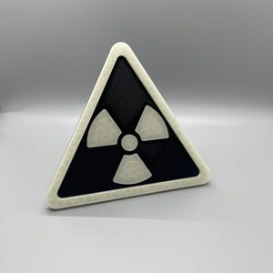 May include: A black and white triangular warning sign with a white radiation symbol inside. The sign has a white border.