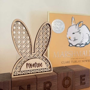 May include: A wooden bunny ear decoration with the name "Monroe" cut out. The bunny ears have a woven pattern. The decoration sits on top of wooden blocks spelling out "Monroe". A children's book with a rabbit illustration is in the background.