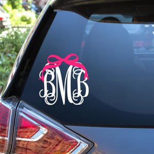May include: White monogram decal with the letters BMB, adorned with a pink bow. The decal is applied to a car window. The words "Sassy Monograms" are printed on the side of the window.