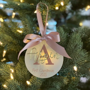May include: A circular Christmas ornament with the name "Aspen" and "First Christmas 2023" in a script font. It features a pink ribbon and bow for hanging. The background is a Christmas tree with fairy lights.
