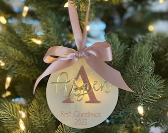 Könnte beinhalten: Runder Weihnachtsschmuck mit dem Namen "Aspen" und "First Christmas 2023" in Schreibschrift. Er hat ein rosa Band und eine Schleife zum Aufhängen. Der Hintergrund ist ein Weihnachtsbaum mit Lichtern.