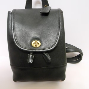 May include: Black leather backpack with a flap closure and a gold-tone clasp. The backpack has a top handle and adjustable shoulder straps. A small leather tag hangs from the top. The bag is a classic design.