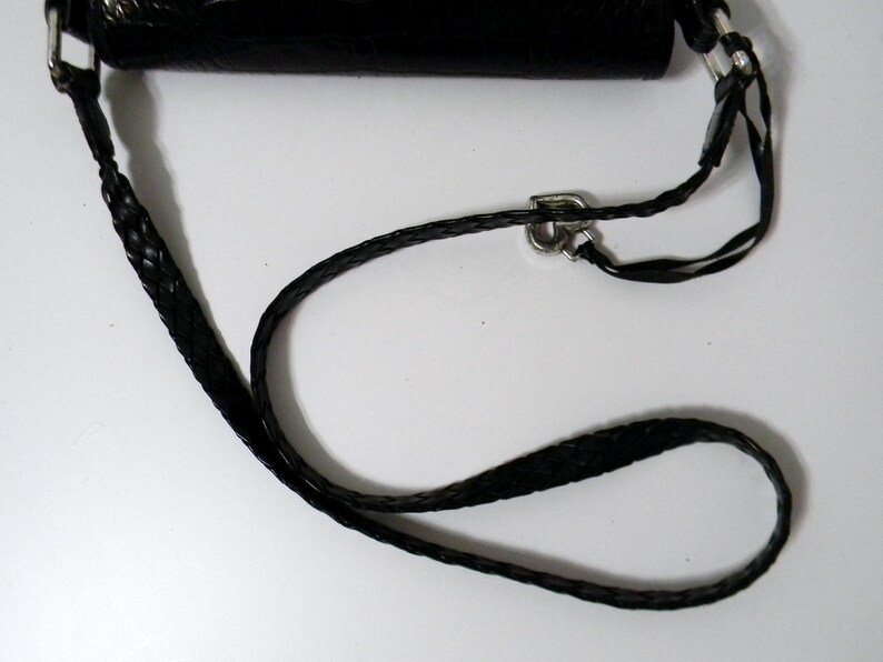 Może przedstawiać: Czarna sk&oacute;rzana torba na ramię z plecionym paskiem i srebrnymi elementami. Pasek jest regulowany i wydaje się być wykonany z wytrzymałego materiału. Torba jest częściowo widoczna, ukazując teksturowaną powierzchnię.