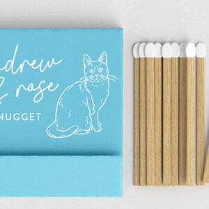 May include: A light blue matchbox with the words "andrew & rose + nugget" and a white cat illustration. Several wooden matches with white tips are next to the matchbox. The matches are made of wood.
