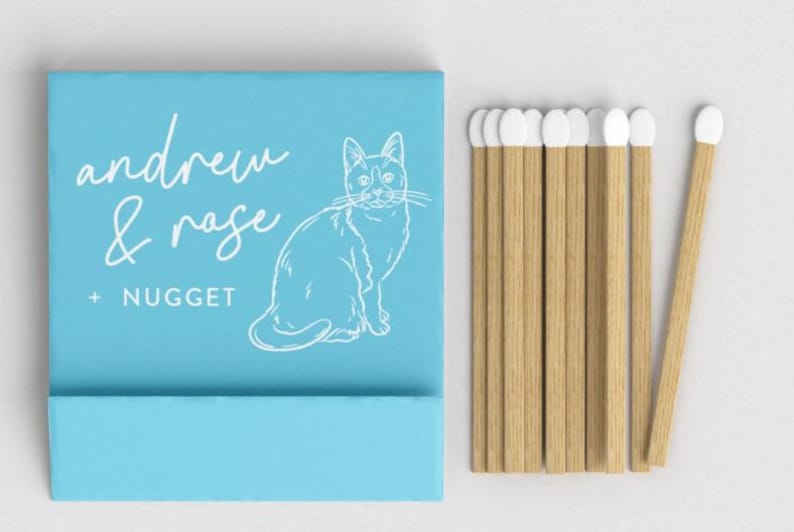 May include: A light blue matchbox with the words "andrew & rose + nugget" and a white cat illustration. Several wooden matches with white tips are next to the matchbox. The matches are made of wood.