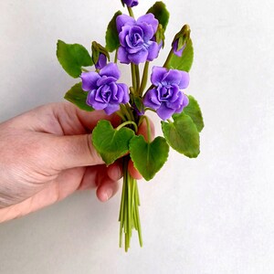 Peut inclure: Un bouquet de fleurs violettes avec des feuilles vertes. Les fleurs sont en pâte polymère et ressemblent à des violettes.