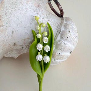 May include: A handcrafted lily of the valley brooch. The brooch features white bell-shaped flowers, green leaves, and a green stem. The brooch is attached to a white, textured surface with a brown ring.