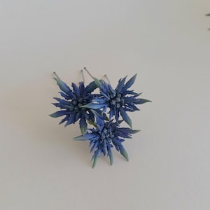 Puede incluir: Tres horquillas florales azules con pétalos puntiagudos y puntas verdes. Las flores están dispuestas en un grupo, con las horquillas que se extienden desde la parte posterior. El fondo es una superficie blanca lisa.