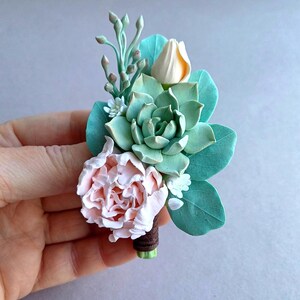 May include: A small bouquet of artificial flowers, featuring a pink peony, a white succulent, and a light yellow flower. The bouquet is tied with brown twine and has green leaves.