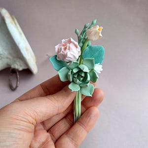 May include: A small bouquet of faux flowers made of polymer clay. The bouquet features a pink rose, a white flower, and a succulent. The flowers are attached to a wire stem.