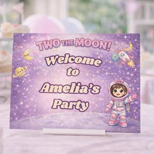 以下が含まれることがあります： 「TWO THE MOON！Welcome to Amelia's Party」の文字と、月、惑星、ロケット、宇宙飛行士のイラストが描かれた紫色のパーティーサイン。カップケーキと飲み物が背景にあるテーブルにサインが置かれています。