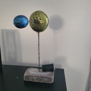 May include: A metal sculpture with a blue and gold sphere on a metal rod. The sculpture is sitting on a concrete block.