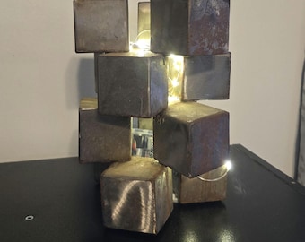 May include: A sculpture made of stacked metal cubes with a string of fairy lights woven through the gaps. The cubes are a dark gray color with a rusty patina.