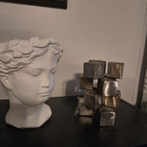 May include: A white plaster bust of a woman with a laurel wreath on her head, next to a sculpture made of stacked metal cubes.