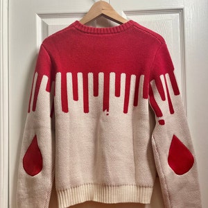 May include: A cream coloured sweater with red dripping paint design. The sweater has red elbow patches in the shape of a teardrop.
