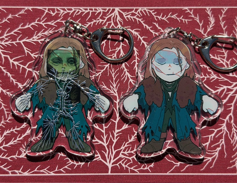 May include: Two acrylic keychains with a cartoon zombie design. The zombie has green skin, brown hair, and a teal coat. Each keychain has a silver-coloured metal ring for attaching to keys or bags.