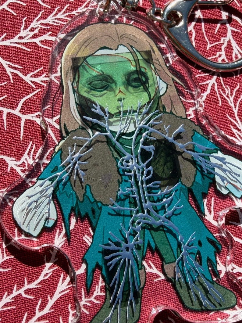 May include: A clear acrylic keychain with a zombie illustration. The zombie has green skin, brown hair, and teal clothing. White, branch-like details overlay the figure. A silver keyring is attached.