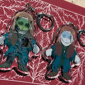 May include: Two acrylic keychains depicting zombie-like figures with green and blue skin, brown hair, and torn attire. Each keychain has a silver clasp. The background is a red surface with a white, branch-like pattern.