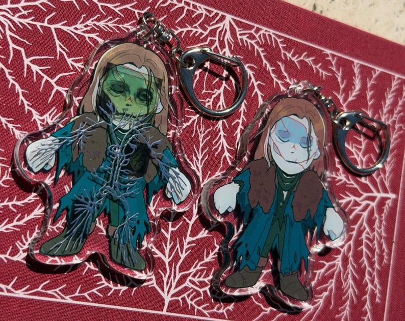May include: Two acrylic keychains depicting zombie-like figures with green and blue skin, brown hair, and torn attire. Each keychain has a silver clasp. The background is a red surface with a white, branch-like pattern.