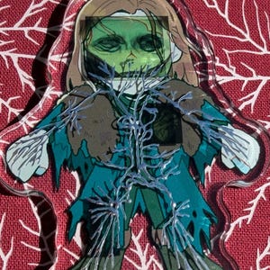May include: A clear acrylic charm with a zombie illustration. The zombie has green skin, brown hair, and teal clothing. Silver vein-like details overlay the design. The charm is on a red background with white branch patterns.