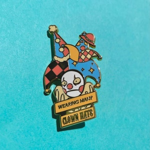 May include: A gold-colored enamel pin featuring a cartoon jester wearing multiple colorful hats. The jester has a white face with red lips and black eyes. The pin has a yellow banner with the text "WEARING MANY CLOWN HATS".