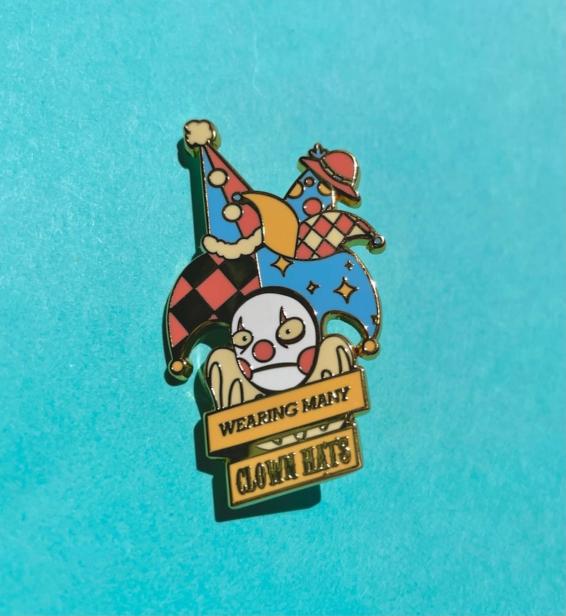 May include: A gold-colored enamel pin featuring a cartoon jester wearing multiple colorful hats. The jester has a white face with red lips and black eyes. The pin has a yellow banner with the text "WEARING MANY CLOWN HATS".