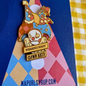 May include: Gold enamel pin featuring a cartoon character wearing multiple hats. The hats are a jester's hat, a party hat, and a top hat. The character is holding a sign that says "Wearing Many Clown Hats".