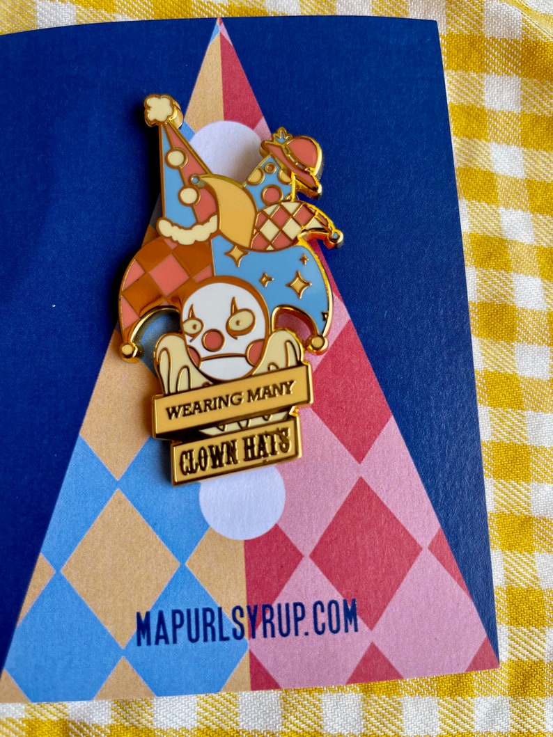 May include: Gold enamel pin featuring a cartoon character wearing multiple hats. The hats are a jester's hat, a party hat, and a top hat. The character is holding a sign that says "Wearing Many Clown Hats".