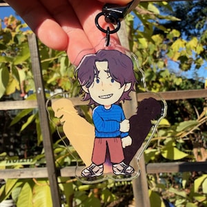 Könnte beinhalten: Ein klarer Acryl-Schlüsselanhänger mit einer Cartoon-Illustration einer Person mit lila Haaren, einem blauen Pullover und einer braunen Hose. Der Schlüsselanhänger hat einen schwarzen Verschluss und einen schwarzen Ring. Der Hintergrund ist eine sonnige Szene im Freien.