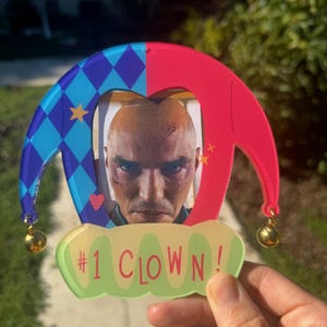 Könnte beinhalten: Ein farbenfroher Narrenhut-förmiger Ornament mit einem Foto eines Mannes. Der Hut ist blau und rot mit goldenen Glöckchen. Der Text "#1 CLOWN!" befindet sich auf einem grünen, wolkenförmigen Banner.