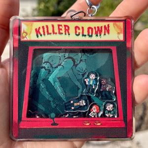 May include: A red and black arcade-style keychain with the text "Killer Clown" on the top. The keychain features a pixelated image of a green forest with several characters in the foreground.