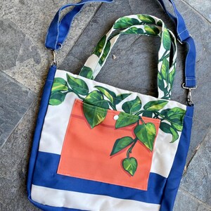 Puede incluir: Un bolso tote con una base a rayas azules y blancas y una correa azul. El bolso presenta un fondo blanco con un estampado de hojas verdes y un bolsillo naranja. El bolso tiene una bandolera y un asa.
