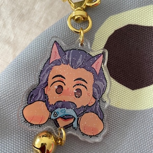 Puede incluir: Un llavero dorado con un dije transparente que presenta un personaje de dibujos animados con cabello morado, barba y orejas de gato. El personaje sostiene un pez azul en su boca. El dije tiene una campana dorada.