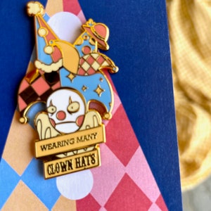 May include: Gold enamel pin featuring a white cartoon character wearing multiple colorful hats. The character is holding a sign that reads "Wearing Many Clown Hats".